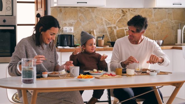 Smiling parents and little girl talking at table with tasty fruit slices and hot drinks in house kitchen