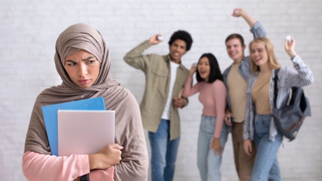 Peer problems, victim, racism, humiliation and bullying in college, school or university. Multiracial young students laugh and throw paper at upset arabian woman with books, studio shot, empty space