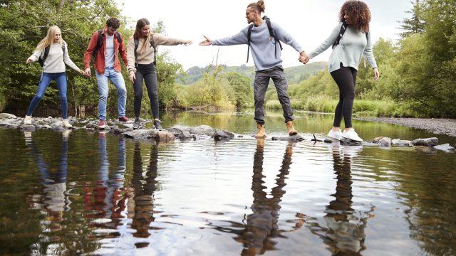 Young adult friends reaching to help each other cross a stream balancing on stones during a hike